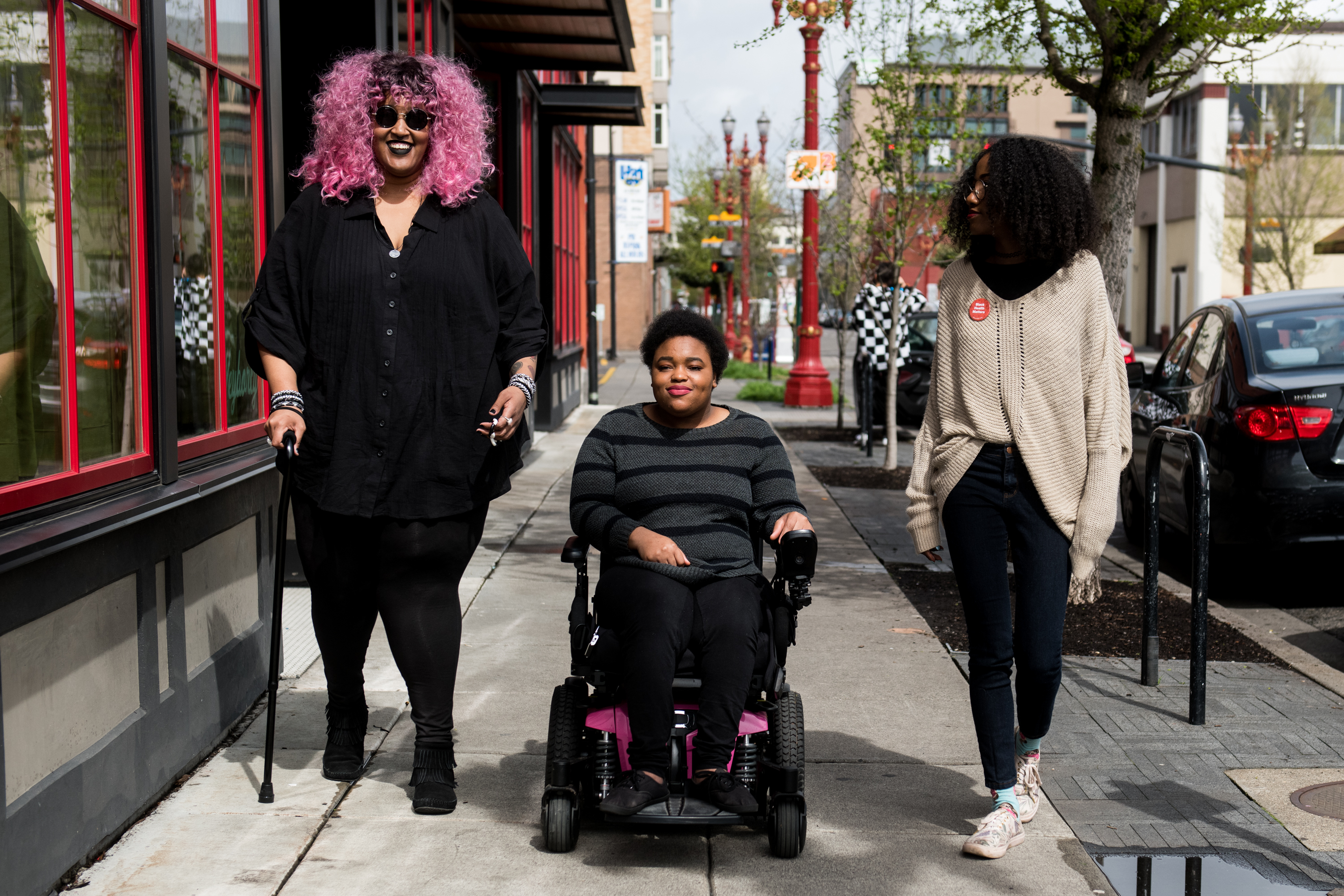 The 15-Minute City Has an Imaginary Resident Photograph of three disabled Black friends moving along a city sidewalk, including a person using a power wheelchair and another using a cane.