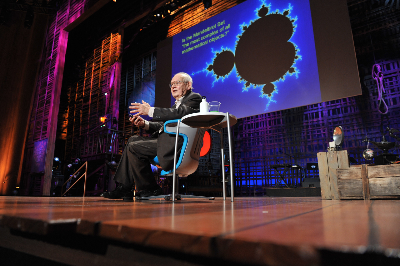 Benoit Mandelbrot speaking on the TED stage with a large visual screen behind him.